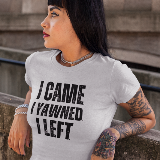 Woman wearing a white t-shirt with 'I CAME I YAWNEd I LEFT' text, leaning against a stone wall.