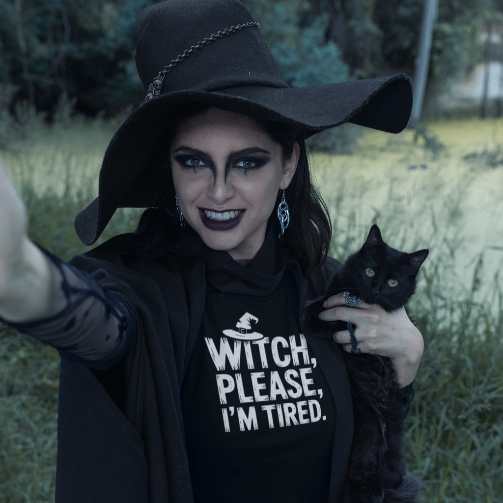 Person wearing a black witch hat and 'Witch, Please, I'm Tired' shirt holding a black cat outdoors.
