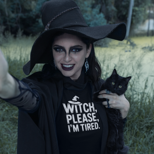 Person wearing a black witch hat and 'Witch, Please, I'm Tired' shirt holding a black cat outdoors.